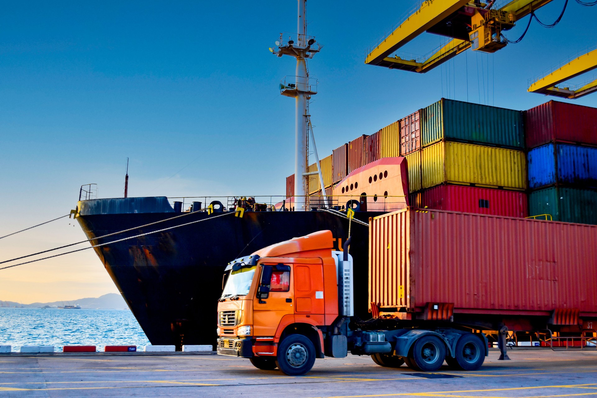 Truck carrying forty-foot container leaving port terminal with ship and quay crane on the background. Seaport operation activities, container shipping, and logistics.